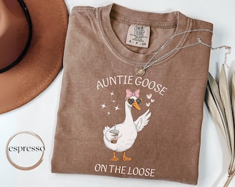 May include: Brown Comfort Colors t-shirt with the phrase "Auntie Goose on the Loose" and a cartoon goose wearing sunglasses and a bow. A silver necklace and a brown hat are also visible.