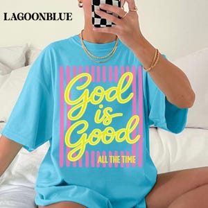 May include: A bright blue short-sleeved t-shirt featuring a graphic design with vertical pink stripes and the yellow cursive text God is Good, with the additional text ALL THE TIME printed in yellow block letters at the bottom of the design.