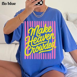 May include: A periwinkle blue short-sleeved t-shirt featuring a graphic design with vertical pink stripes and the yellow cursive text "Make Heaven Crowded" centered on the front. The shirt has a crew neckline and a relaxed, loose-fitting silhouette.