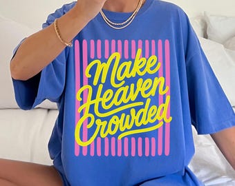 May include: A periwinkle blue short-sleeved t-shirt featuring a graphic design with vertical pink stripes and the yellow cursive text "Make Heaven Crowded" centered on the front. The shirt has a crew neckline and a relaxed, loose-fitting silhouette.