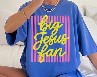 May include: A flo blue short-sleeve t-shirt featuring a graphic design with vertical pink stripes and the yellow cursive text "big Jesus fan" centered on the front. The shirt has a crew neckline and a loose, relaxed fit.