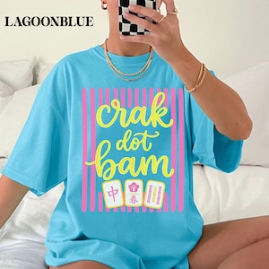 May include: A bright blue short-sleeve t-shirt featuring a graphic print with vertical pink stripes and the yellow cursive text "crak dot bam" above three mahjong tiles displaying a red character, a pink flower, and three green bamboo sticks.