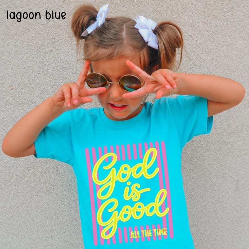 May include: A lagoon blue short-sleeved t-shirt featuring a graphic design with vertical pink stripes and the yellow text "God is Good ALL THE TIME" printed on the front. The shirt is made of a solid lagoon blue fabric with a crew neckline.
