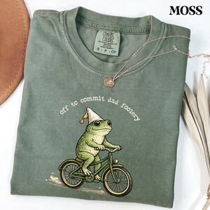 May include: Moss green t-shirt with a graphic of a frog riding a bicycle and wearing a party hat. The text "off to commit dad foolery" is printed above the frog. A silver necklace with a pendant is on the shirt.