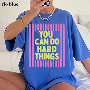 May include: A periwinkle blue short-sleeve t-shirt featuring a graphic design with vertical pink stripes framing the neon yellow text that reads YOU CAN DO HARD THINGS. The shirt has a crew neckline and a loose, relaxed fit for casual wear.