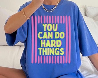 May include: A periwinkle blue short-sleeved t-shirt featuring a graphic design with vertical pink stripes framing the neon yellow text that reads YOU CAN DO HARD THINGS. The shirt has a crew neckline and a loose, relaxed fit for casual wear.
