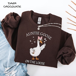 May include: Dark chocolate-colored sweatshirt with the text "AUNTIE GOOSE ON THE LOOSE" and a cartoon goose wearing sunglasses and a pink bow. A silver necklace is draped across the sweatshirt. The words "DARK CHOCOLATE" are in the upper left corner.
