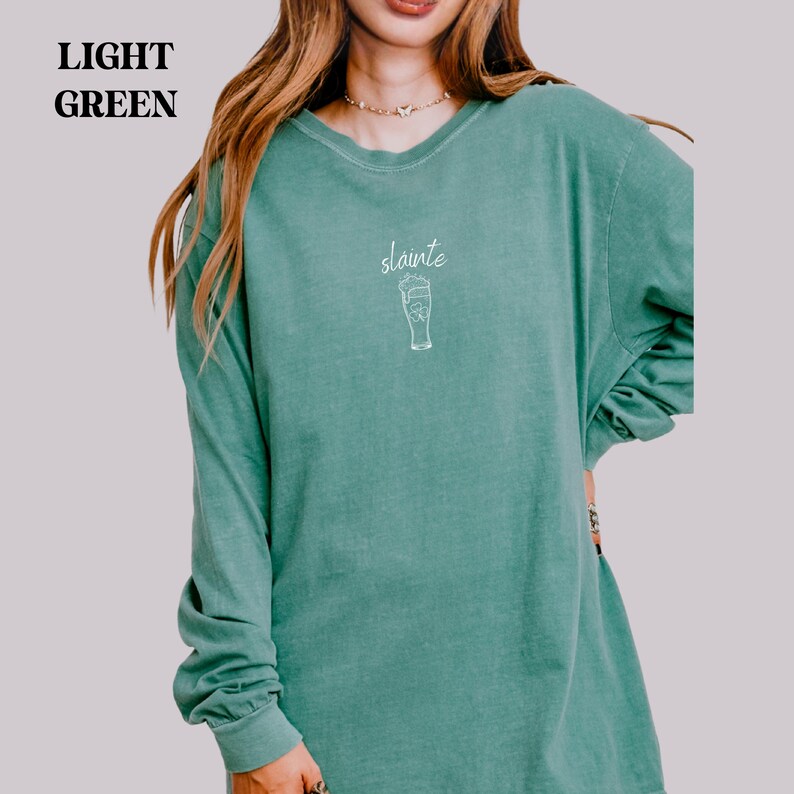 May include: Light green long-sleeve shirt with the word "sl&aacute;inte" and a beer glass graphic in white. The shirt has a crew neck and long sleeves. The shirt is a solid light green color.