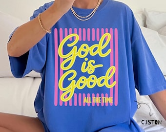 May include: A blue short-sleeved t-shirt featuring a graphic design with yellow cursive text that reads God is Good and smaller yellow text that reads ALL THE TIME, framed by vertical pink stripes on a blue background.
