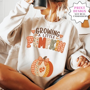 May include: A beige crewneck sweatshirt featuring a printed graphic with the text "GROWING A LITTLE PUMPKIN" in various patterns including plaid, floral, and stripes. Below the text is a large pumpkin illustration with baby footprints.