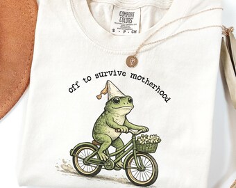 May include: Ivory-colored t-shirt with a whimsical illustration of a frog riding a bicycle, wearing a dunce cap. The text "off to survive motherhood" is arched above the frog. A gold necklace and a brown hat are also visible.