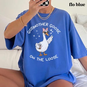 May include: Flo blue t-shirt with a graphic of a goose wearing sunglasses and a bow, holding a coffee cup. The text reads "GODMOTHER GOOSE ON THE LOOSE" in white. The shirt is a solid blue color.