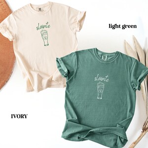 May include: Two t-shirts, one ivory and one light green, are displayed. Each shirt features the word "sl&aacute;inte" above a graphic of a beer glass with a shamrock. The ivory shirt is on the left, and the light green shirt is on the right.