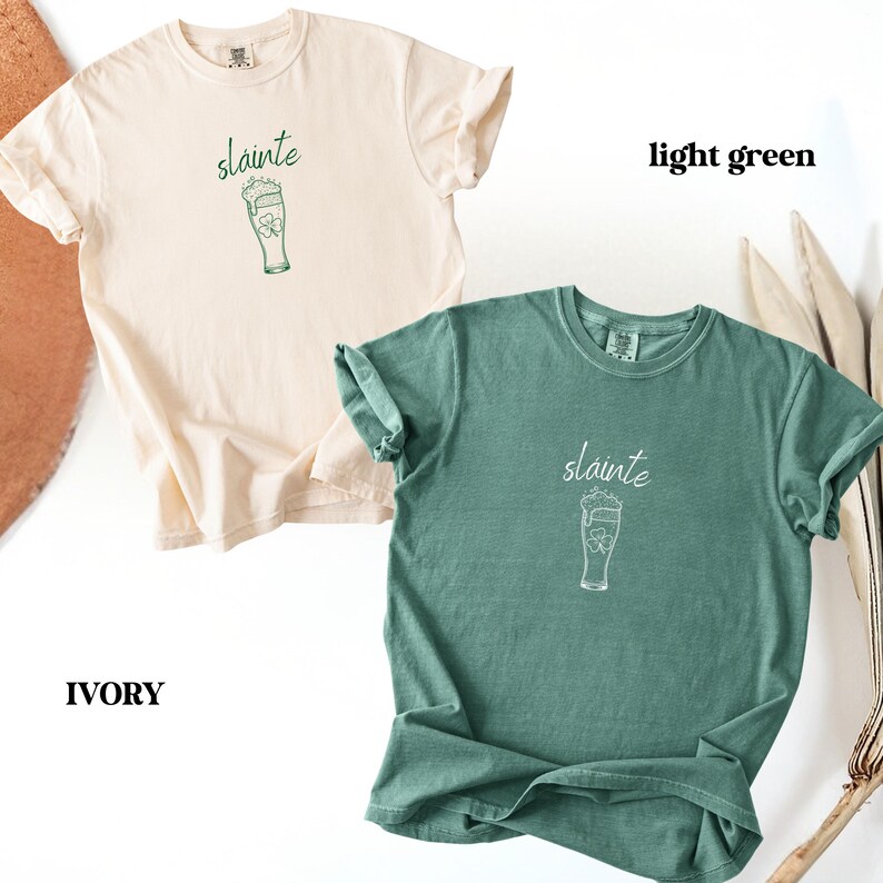 May include: Two t-shirts, one ivory and one light green, are displayed. Each shirt features the word "sl&aacute;inte" above a graphic of a beer glass with a shamrock. The ivory shirt is on the left, and the light green shirt is on the right.