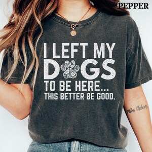 May include: Dark gray t-shirt with the text "I LEFT MY DOGS TO BE HERE... THIS BETTER BE GOOD." The word "DOGS" features a paw print design. The shirt is a comfortable, casual style.
