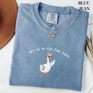 May include: Blue jean colored t-shirt with the text "off to be the fun aunt" and a cartoon goose wearing sunglasses and a pink bow. A gold necklace and the Comfort Colors brand tag are visible.
