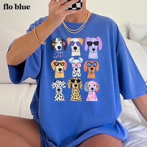 May include: A blue t-shirt featuring a graphic print of nine stylized dog heads arranged in a grid. Each dog wears different colored sunglasses or floral accessories. The text "flo blue" is printed in the upper left corner of the image.