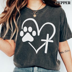 May include: A charcoal gray short-sleeve t-shirt featuring a white graphic of a heart combined with a paw print and a cross. The shirt has a crew neckline and a vintage-washed appearance. The word PEPPER is printed in the top right corner.