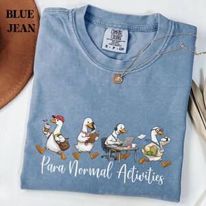 May include: Blue jean Comfort Colors t-shirt with a graphic of four cartoon geese engaged in various activities. The text "Para Normal Activities" is printed below the geese. A gold necklace is draped across the shirt.