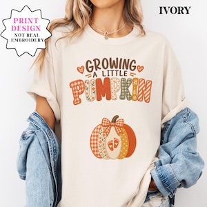 May include: An ivory t-shirt featuring a printed graphic with the text "GROWING A LITTLE PUMPKIN" in brown letters. The design includes a large pumpkin illustration with orange, white, and green plaid and floral patterns, plus two small baby footprints.