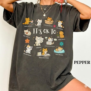 May include: Dark grey t-shirt with the text "IT'S OK TO" and illustrations of cats. The cats are depicted in various scenarios, such as making mistakes, avoiding eye contact, and taking breaks. The word "PEPPER" is printed on the bottom right.