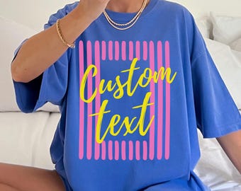 May include: Flo blue t-shirt with a custom text design. The shirt features a rectangular design with pink vertical stripes and the words "Custom Text" in yellow. The text "flo blue" is in the upper right corner.
