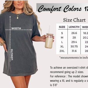 May include: A woman wearing a dark gray oversized t-shirt dress. The image includes a size chart with measurements in inches, and the text "Comfort Colors 1717". The chart shows sizes S to 2XL, with length and width measurements.