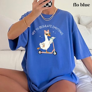 May include: Flo blue t-shirt with a graphic of a goose wearing sunglasses and a bow riding a pink scooter. The text "OFF TO REGULATE EMOTIONS" is arched above the goose. The shirt is a comfortable, oversized fit.
