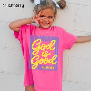May include: A bright pink short-sleeved t-shirt featuring a graphic print with vertical blue stripes and the yellow text God is Good ALL THE TIME. The shirt has a crew neckline and a relaxed fit, showcasing a bold color contrast between the pink fabric.