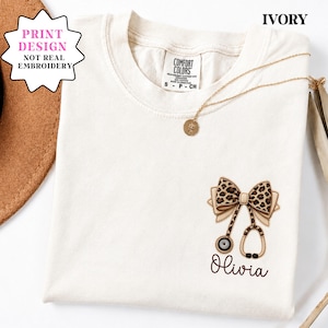 May include: Ivory-colored t-shirt with a leopard print bow and stethoscope design. The design includes the name "Olivia" in script. A gold necklace with a pendant is draped on the shirt. The shirt has a tag that says "Comfort Colors".
