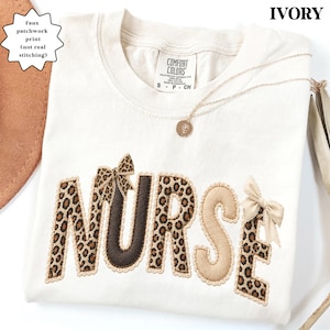 Könnte beinhalten: Elfenbeinfarbenes T-Shirt mit dem Wort "NURSE" im Leopardenmuster. Die Buchstaben sind mit einem gewellten Rand umrandet und mit Schleifen verziert. Eine goldene Halskette mit Anhänger liegt auf dem Shirt.