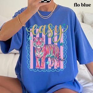 May include: Flo blue t-shirt with a colorful tiger graphic. The tiger has pink, green, and orange stripes, set against a pink and white striped background. The word "easy" is written in gold script above the tiger, with blue stars and a decorative border.