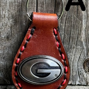 May include: Brown leather keychain with a red and black stitched border. The keychain features a silver metal emblem with a black 'G' inside a white oval.
