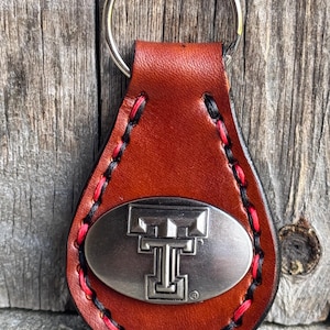 May include: Brown leather keychain with a silver oval charm featuring the Texas Tech University logo. The keychain has red and black stitching.