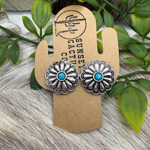 May include: A pair of silver earrings with turquoise stones in the center of each flower-shaped design. The earrings are displayed on a brown card with the text "Sunset Cactus Co."