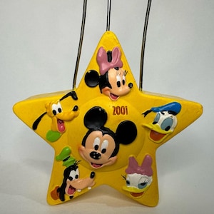 May include: Yellow star-shaped ornament with Mickey Mouse, Minnie Mouse, Donald Duck, Goofy, and Pluto characters. The ornament has a wire hanger and the year 2001 printed on it.