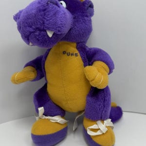 May include: A plush toy dinosaur in purple and gold. The dinosaur has a yellow belly and feet, with purple arms, legs, and head. The word "DUKE" is embroidered on the chest. The dinosaur is wearing white ribbon shoes.
