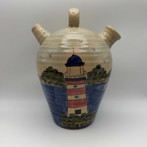 May include: A beige ceramic pitcher with a handle and two spouts, featuring a hand-painted scene of a red and white striped lighthouse on a rocky shore with blue water, green foliage, and small black birds flying in the sky above the structure.