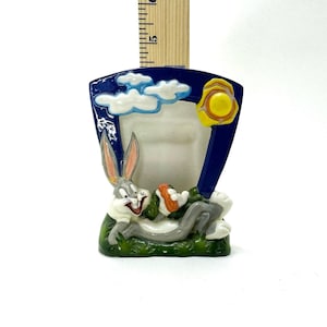 May include: A ceramic picture frame featuring Bugs Bunny lying in green grass while holding a carrot. The frame has a dark blue background with white clouds and a yellow sun. A wooden ruler shows the frame is approximately 5 inches tall.
