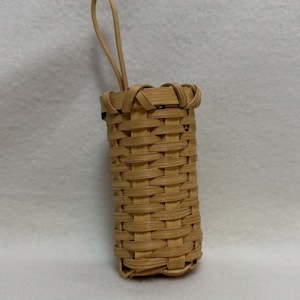 May include: A small, cylindrical woven basket made of light tan natural reeds. The basket features a tight, uniform weave pattern throughout its body and includes a single, thin, flexible loop handle extending upward from the top rim.