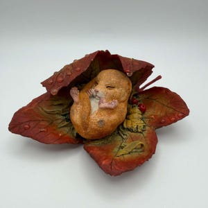 May include: A small, light brown, sleeping mouse figurine curled up inside a large, textured, deep red and orange autumn leaf. The leaf features visible veins, small water droplets on its surface, and a cluster of tiny red berries near the mouse.