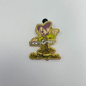 May include: A metal enamel pin featuring the character Grumpy from Snow White, wearing a purple hat and olive green tunic. The text I'M WITH GRUMPY is printed in white, and the text WALT DISNEY WORLD is printed in gold at the bottom of the pin.