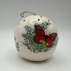 May include: A spherical white ceramic ornament featuring a painted monarch butterfly with orange and black wings resting on green foliage with small blue flowers. The top of the ornament has several small circular holes and a gold hanging cord.