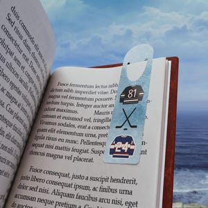 Può includere: Un segnalibro a tema hockey con disegni di maglie con i numeri 81 e 24, insieme a bastoni da hockey incrociati. Il segnalibro è azzurro ed è posto all'interno di un libro aperto.