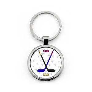 May include: Silver-colored keychain with a round pendant featuring two crossed hockey sticks. The sticks are black, blue, and red, with the numbers "1410" and "1221" above and below. The background has a pattern of small gray hearts.