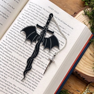May include: A black leather bookmark featuring a dragon and sword design. The bookmark is attached to a silver chain with a cross charm. The bookmark is lying open on a book with the text "FAIRY TALE" visible.