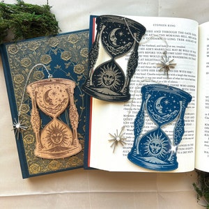 May include: Two wooden hourglass bookmarks with a sun and moon design. The bookmarks are hanging from a silver chain with a star charm. The bookmarks are on top of a book with the text "Here and through the gate God..." printed on the page.