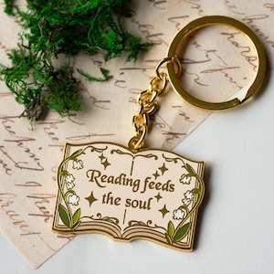 Może przedstawiać: Złoty brelok do kluczy z zawieszką w kształcie otwartej książki. Zawieszka zawiera napis "Reading feeds the soul" i jest ozdobiona elementami kwiatowymi. Brelok jest przymocowany do złotego łańcuszka i kółka.