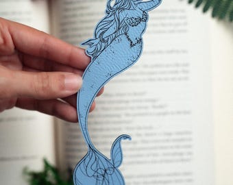 May include: A light blue mermaid bookmark with black outline details. The mermaid has long flowing hair and a patterned top. The bookmark is held in front of an open book, with a blurred background of greenery and a flower.