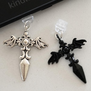 May include: Two cross-shaped charms with bat wing designs. One is silver-toned, the other is black. Both have a clear plastic clip and a small metal ring. The word "kindle" is visible on a black surface in the background.